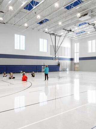 A spacious gymnasium features bright lighting, a basketball hoop, and a few individuals seated on the polished floor.