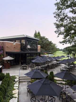 A modern restaurant with a spacious outdoor patio, featuring black umbrellas over tables, surrounded by greenery and a serene landscape.