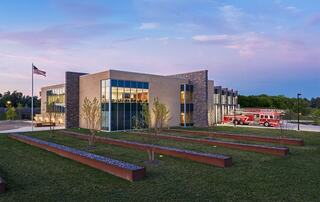 A modern building with large windows and a landscaped lawn is adjacent to a fire truck, under a colorful sky at dusk. The American flag flutters.
