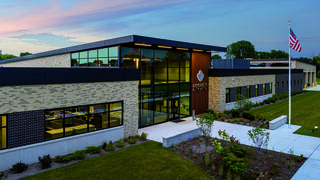 A modern brick building with large windows, landscaped grounds, and an American flag, set against a colorful sunset sky.