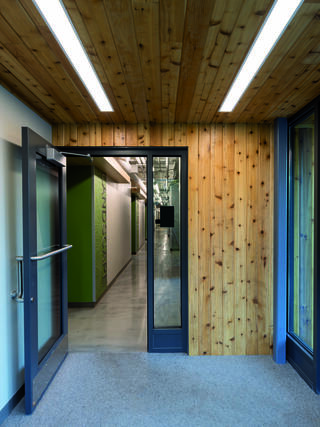 A wooden entrance features glass doors leading to a well-lit corridor with green accents and sleek flooring. Modern design elements are evident.