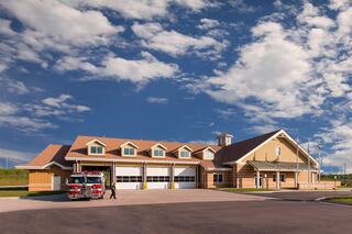 A spacious fire station with a red fire truck parked outside, set against a backdrop of blue skies and scattered clouds.