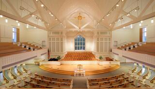A spacious, well-lit sanctuary featuring wooden pews, a central stage, and large windows, creating a serene and inviting atmosphere.