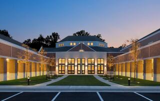 A modern brick building with large illuminated entrance doors, surrounded by neatly landscaped greenery and paths, under a twilight sky.