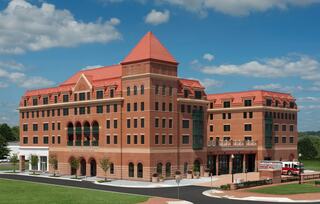 A large brick building with a red-tiled roof features multiple stories, arched windows, and modern design elements, set against a blue sky.