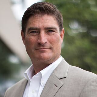 A confident man in a light gray suit and white shirt stands outdoors, with a blurred green background, presenting a professional demeanor.