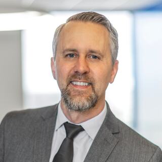 A confident professional with a light beard and neatly styled hair, dressed in a gray suit and tie, smiles warmly in a modern office setting.