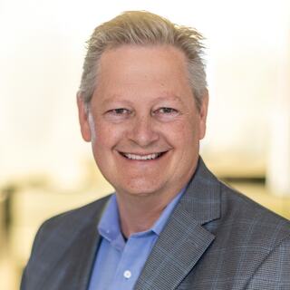 A smiling man with short, light hair wears a gray blazer over a blue shirt, exuding a professional and approachable demeanor.