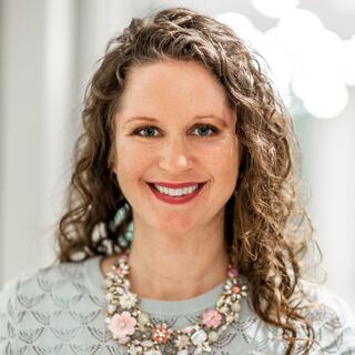 A smiling woman with curly hair wears a light sweater and a colorful, floral statement necklace, exuding warmth and confidence.