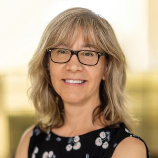 A woman with shoulder-length, wavy hair and glasses smiles warmly, wearing a floral dress, against a softly blurred background.