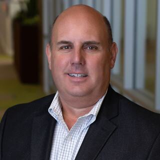 A middle-aged man with short hair, wearing a dark blazer over a checked shirt, smiles warmly against a softly blurred background.