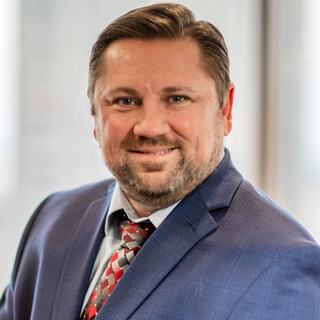 A smiling man in a navy suit and patterned tie stands against a bright background, exuding confidence and professionalism.