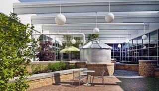 A modern courtyard features green plants, a round metal structure, and stylish outdoor seating under hanging lights and an umbrella.