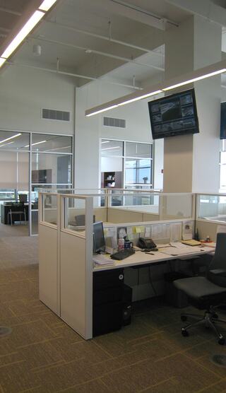 A modern office space featuring cubicles, a desk with a computer, and a wall-mounted TV, illuminated by bright overhead lights.