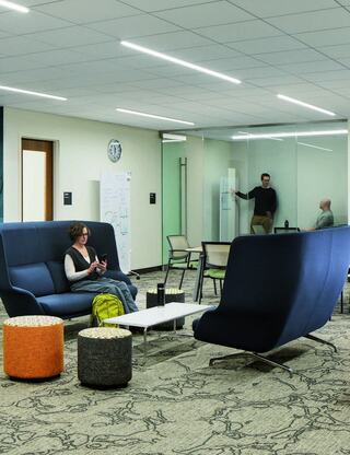 A modern lounge with stylish seating, colorful poufs, and a glass-walled meeting area. People are engaged in discussions and working.