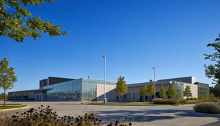 A modern building with large glass windows is surrounded by greenery and blue skies, showcasing a sleek architectural design.