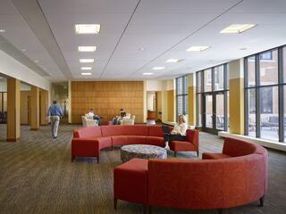 A spacious, modern lounge features circular red seating, large windows, and visitors engaged in conversation or reading, creating a welcoming atmosphere.