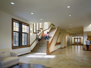 A modern, well-lit lobby features a staircase where three people sit, surrounded by large windows and a welcoming atmosphere.