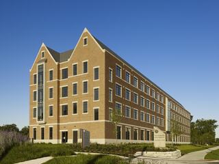 A modern brick building with a distinctive roofline, featuring multiple windows and landscaped surroundings under a clear blue sky.