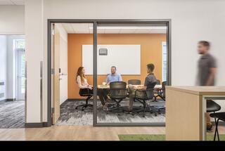 A modern conference room showcases four people engaged in discussion, with a bright orange accent wall and contemporary furnishings.