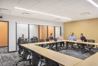 A modern meeting room features a long table with four people discussing while another person works independently in the background.