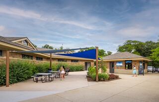 A modern building with outdoor seating under a blue shade sail, surrounded by greenery. People enjoy the space in a welcoming atmosphere.