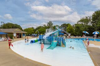 A vibrant pool area features a large water slide, sunbathers, and children enjoying the water under a partly cloudy sky. Lifeguards are on duty.