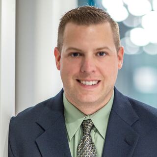 A professional man smiles confidently, dressed in a dark suit, light green shirt, and patterned tie, against a softly blurred background.