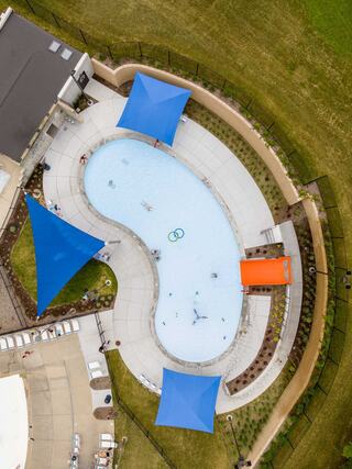 Aerial view of a curved swimming pool surrounded by green grass, featuring blue shade structures and orange slides, with some people swimming.