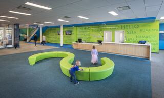 A modern space features a vibrant green seating area, with children playing and colorful greetings in multiple languages on the walls.