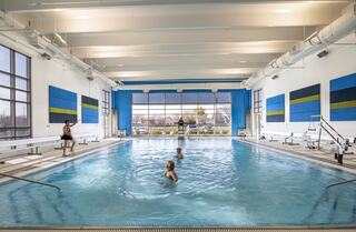 A bright indoor swimming pool features children playing, a lifeguard nearby, and colorful wall panels, with large windows letting in natural light.