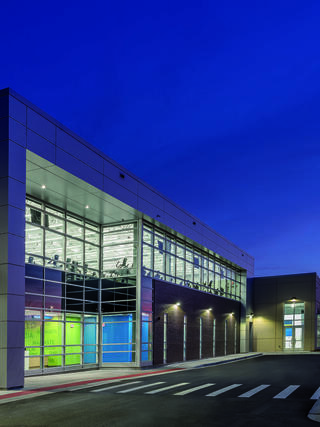A modern building features large glass windows, showcasing a bright interior with colorful accents, set against a twilight sky.