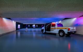 A dimly lit shooting range features a police vehicle with flashing lights, while a lone figure stands at the far end amidst colorful lighting.