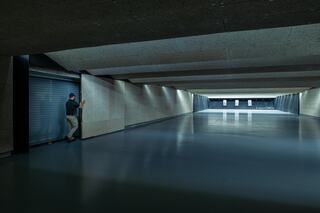 A man stands near a large, open space with smooth floors and walls, leading to a distant lit area with targets. The environment appears modern and structured.