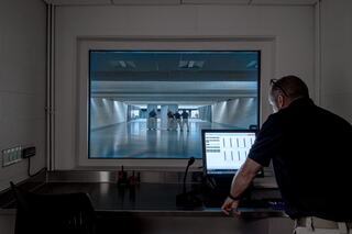 A person stands at a workstation, monitoring a spacious, brightly lit area where several individuals are gathered.