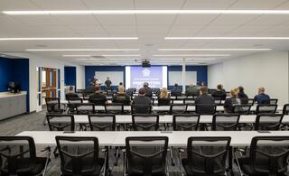 A training session takes place in a modern classroom with attendees seated, facing a screen displaying a law enforcement logo.
