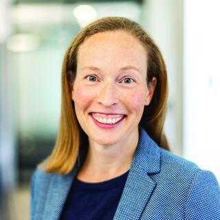 A smiling woman with straight, shoulder-length hair wears a blue blazer over a dark top, set in a modern, softly blurred environment.