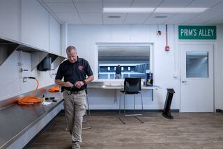 A person in a uniform stands in a modern room with stainless steel surfaces and equipment, focused on a task, with a sign reading "PRIM'S ALLEY."