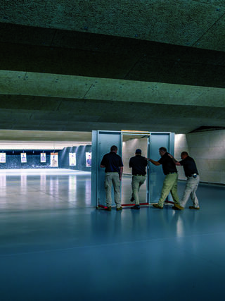 Four individuals move a shooting target setup in a well-lit shooting range, preparing for practice against distant targets.