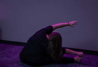 A person stretches on the floor in a dimly lit room, with purple lighting and illuminated squares on the wall creating a serene atmosphere.