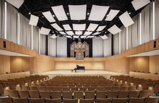 A spacious concert hall with wooden interiors, featuring a grand organ, a grand piano on stage, and neatly arranged seating for an audience.