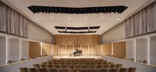 A spacious auditorium features wooden acoustics, a grand piano on stage, and rows of empty chairs, all elegantly designed for performance.