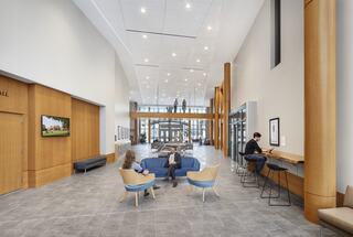 A modern, spacious lobby featuring wooden accents, seating areas, and people working or relaxing, with large windows allowing natural light.