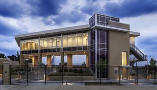 A modern, multi-level building with large glass windows, surrounded by greenery, features a staircase and a welcoming design under a cloudy sky.