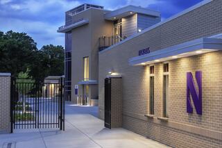 A modern, sleek building with purple accents and a gated entrance, set against a twilight sky and surrounded by greenery.