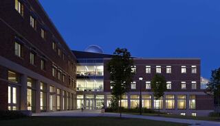 A modern brick building illuminated at dusk, showcasing large glass windows and greenery, creating a welcoming atmosphere.