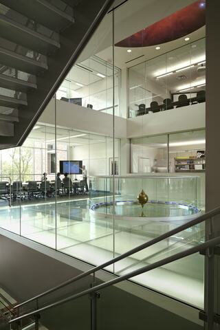 A modern multi-level space features glass walls, an open office area, and a central circular exhibit with a pendulum. Natural light floods in.