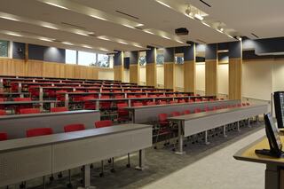 A spacious lecture hall featuring tiered seating with red chairs, modern lighting, and multiple windows providing natural light.