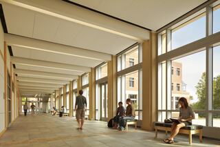 A bright, modern hallway features students sitting and walking, large windows allowing natural light to flood the space, fostering an inviting atmosphere.