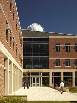 A modern brick building features large glass windows and a distinct dome, with people walking through a paved courtyard under a clear blue sky.